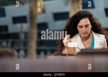 Frau mit langen lockigen Haaren und kastanienbraunen Nägeln nimmt eine Zigarette und Kaffeepause vor einem Café. Weibchen gibt einen Hauch von Rauch ab. Goldener Tag im Freien. Stockfoto