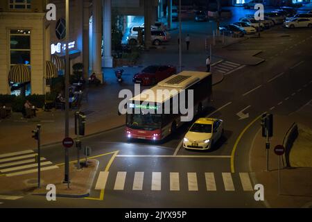 Dubai Bus und Taxi halten an einer Ampel an einem späten Abend in der Innenstadt. Der Fußgängerüberweg und die Insel sind leer. Nur wenige Leute liegen an Tischen und Stühlen vor einem Café. Stockfoto