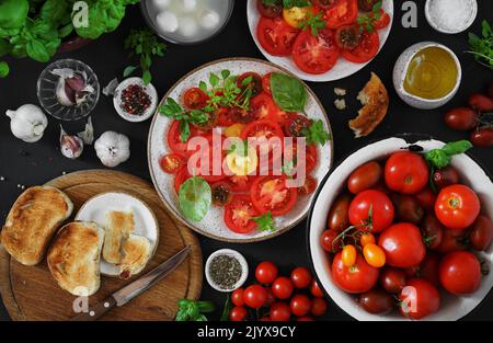 Ganze und in Scheiben geschnittene Bio-Tomaten . Rohe frische Tomaten mit Meersalz und Basilikum und Oliven auf Tellern auf dunklem Hintergrund, Draufsicht Stockfoto