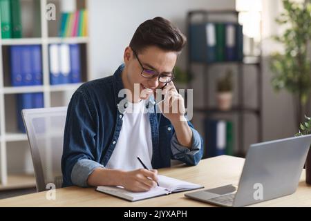 Junger Büroleiter, Der Auf Dem Mobiltelefon Spricht Und Notizen Zu Notepad Nimmt Stockfoto