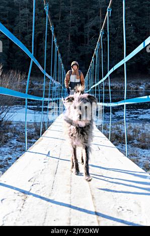 Junger Mann mit Mischlingshund flauschig auf Holzbrücke über Fluss mit ersten Schnee und Eis im Herbst oder frühen Winter Wandern und traveli bedeckt Stockfoto