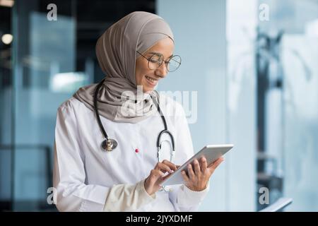 Ärztin in Hijab arbeitet in einem modernen Klinikbüro, muslimische Ärztin verwendet Tablet-Computer, Krankenschwester im medizinischen weißen Mantel und Brille mit Stethoskop. Stockfoto