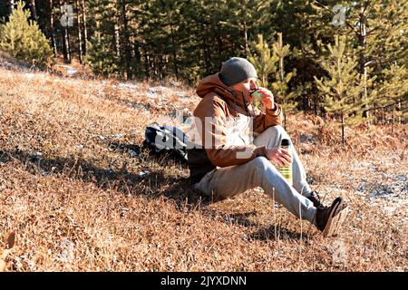 Junger Mann in Herbstkleidung, der Tee aus einer Thermoskanne trinkt, während er am Waldrand in der Abendsonne bei einer Wanderung sitzt und die Natur genießt Stockfoto