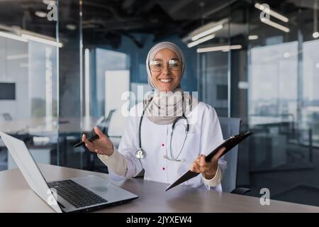 Porträt einer erfolgreichen muslimischen Ärztin in Hijab, einer arabischen Frau, die in einem modernen Klinikbüro arbeitet, lächelt und die Kamera anschaut, einer Ärztin in einer Brille und einem weißen medizinischen Mantel mit Stethoskop. Stockfoto
