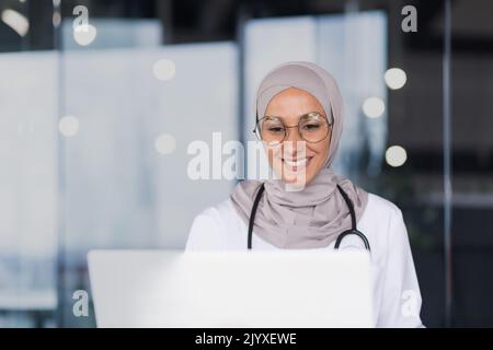 Nahaufnahme Porträt einer Ärztin in einem Hijab arbeitet eine muslimische Frau im Büro einer modernen Klinik, eine Ärztin in einem Kleid und einer Brille benutzt einen Laptop. Stockfoto