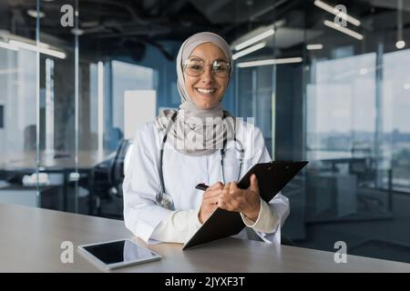 Porträt einer erfolgreichen muslimischen Ärztin in Hijab, einer arabischen Frau, die in einem modernen Klinikbüro arbeitet, lächelt und die Kamera anschaut, einer Ärztin in einer Brille und einem weißen medizinischen Mantel mit Stethoskop. Stockfoto