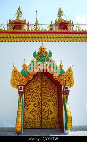 Das prunkvolle Tor von Ucosot des Wat Chong Kham Tempels mit einer Wandskulptur von Garuda oben am Eingang und zwei Statuen von Naga-Schlangen von beiden Seiten Stockfoto