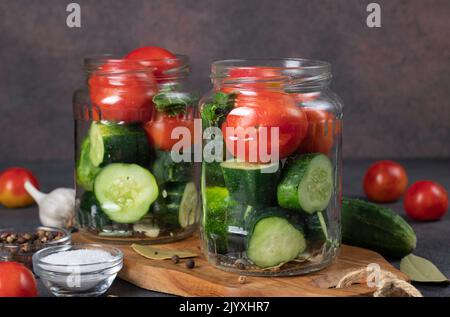 Gurken und Kirschtomaten werden in Gläser mit Knoblauch, Lorbeerblättern, Pfeffer und Dill gegeben. Vorbereitung zum Beizen, Schritt für Schritt Stockfoto