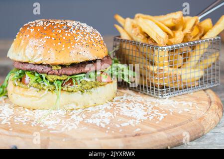 Vegane Burger-Speisekarte mit Pommes auf dem Holzschneidetisch. Hamburger mit veganem Käse, Pesto und Fleischersatz, Rucola, Kirschtomaten. Stockfoto