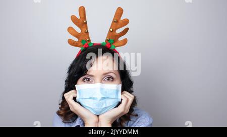 Frau in einer Maske und Hirschhörner Stirnband. Witziges Weihnachtszubehör. Medizinische Maske, Nahaufnahme, selektiver Fokus, Prävention von Pandemie. Stockfoto