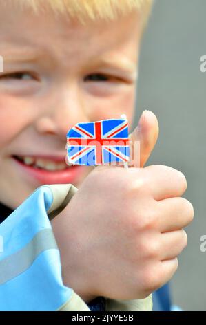 Der blonde Bursche hält die Union Jack-Flagge aus Papier, sein lächelndes Gesicht ist im Hintergrund unscharf. Stockfoto