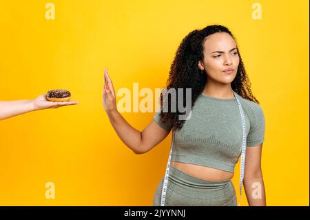 Selbstbewusste sportliche junge schöne hispanische oder brasilianische Frau, stehen auf isoliertem orangefarbenem Hintergrund, macht eine Weigerungsgeste, hält sich an eine gesunde Ernährung, um fit zu sein, gekleidet in ein Sportoutfit. Gesundes Lebensmittelkonzept Stockfoto