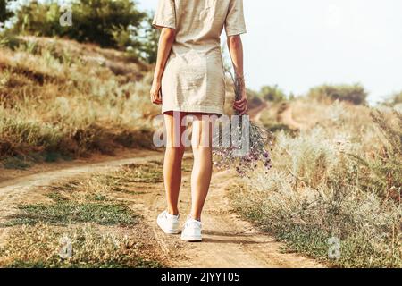 Ein Mädchen mit einem Blumenstrauß aus Herbstblumen geht mit dem Rücken zur Kamera auf einer herbstlichen Straße entlang. Herbstlandschaft Stockfoto