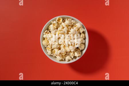 Popcorn in einer weißen Schüssel auf rotem Hintergrund. Blick von oben. Stockfoto