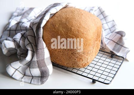 Hausgemachtes Bauernhaus gemischtes Brot. Nahaufnahme von Brot, das in der Backmaschine gebacken wurde. Vollkornbrot auf Trockengestell mit gestreifem Handtuch auf weißem Tisch Stockfoto