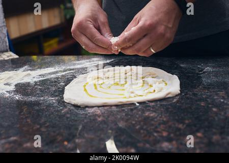 Käse wird mit Sauce auf den Pizzateig gelegt. Der Prozess der Herstellung von hausgemachter Pizza. Das Konzept der hausgemachten Fast Food. Vorderansicht. Stockfoto