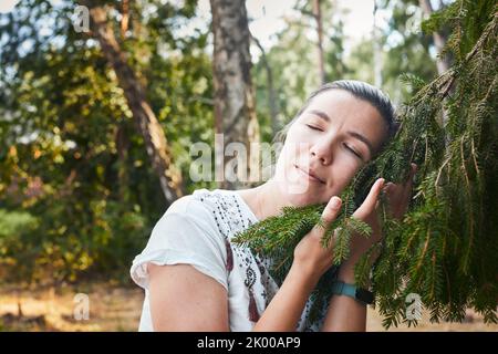 Eine Frau umarmt einen Nadelbaum im Wald. Das Konzept des Lebens in der Nähe von Natur und Wald. Vorderansicht. Stockfoto