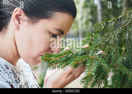 Eine Frau atmet den Duft eines Nadelbaums im Wald ein. Konzept des Lebens in der Nähe von Natur und Wald. Vorderansicht. Stockfoto