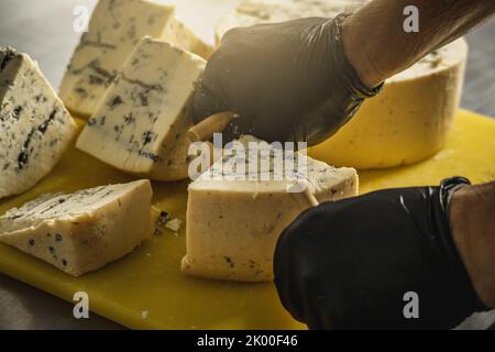 Ein Landwirt in schwarzen Handschuhen schneidet mit einem Slicer einen Kopf aus würzigem Gorgonzola-Käse mit blauem Schimmel in Stücke. Stockfoto