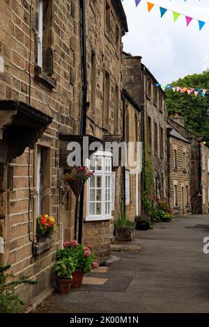 Zwei- und dreistöckige Steinhütten Häuser an der Main Street in Winster Derbyshire, mit Sommerblumen in Türpfannen und Hämmern über der Straße. Stockfoto