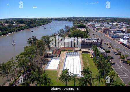 Aus der Vogelperspektive auf eine Küstenstadt mit Fluss, verankerten Booten, Straßen und Gebäuden, die von Bäumen unter blauem Himmel umgeben sind Stockfoto