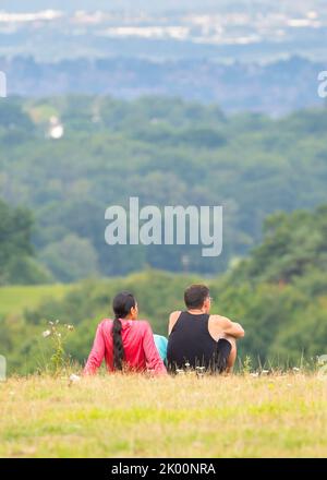 Rückansicht eines Mannes und einer Frau, die isoliert auf einem ländlichen Hügel in Großbritannien sitzen und den Blick auf eine Stadt in der Ferne werfen. Stockfoto