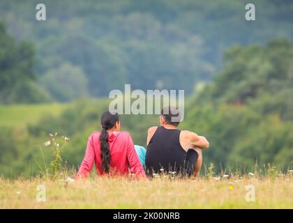 Rückansicht von Mann und Frau, die im Sommer auf einem ländlichen Hügel in Großbritannien sitzen und entspannen und eine Pause von ihrem Spaziergang machen, mit Blick auf die Aussicht. Stockfoto