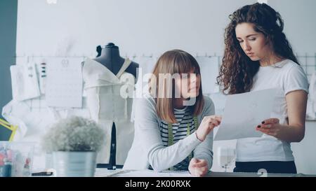 Professionelle Modedesignerinnen schauen sich Skizzen an und diskutieren Details zukünftiger Kleidung. Produktive Zusammenarbeit und Schaffung eines neuen Konzepts für die Bekleidungskollektion. Stockfoto