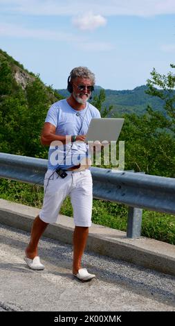 Grauhaariger älterer Mann, diabetischer kranker Freiberufler mit Bart in Sonnenbrille, der an einem Laptop über die Natur und die Berge arbeitete. Ein verrückter und außergewöhnlicher alter Mann, der im Sommer zur Straße geht. Vertikal Stockfoto