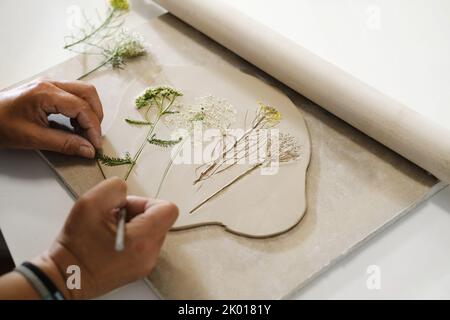 Hands Making Clay handgemachte Keramikplatten, gerollter Ton auf dem Tisch mit Pflanzen und Blumen. Draufsicht auf handgefertigte Keramik, Nahaufnahme Stockfoto