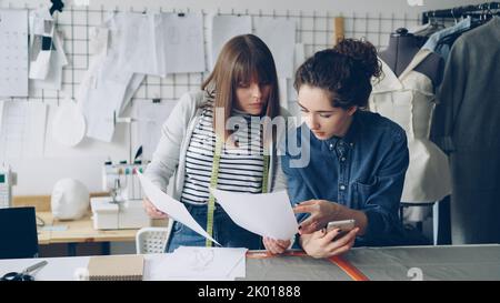 Kreative Modedesignerinnen sind damit beschäftigt, Skizzen in der Nähe des Nähtisches zu sehen. Junge Frauen sprechen und checken Smartphone. Moderne Schneiderei im Hintergrund. Stockfoto