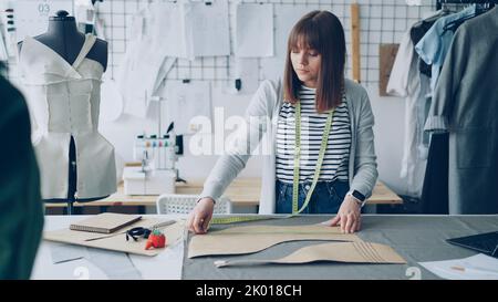 Die junge hübsche Schneiderin misst am Studiotstisch Kleidungsmuster mit Maßband, während sie in ihrem modernen Schneiderladen arbeitet. Vorbereitung auf das Nähkleider-Konzept. Stockfoto