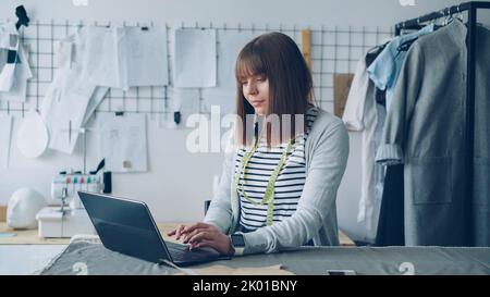 Die junge selbstständige Schneiderin kommuniziert mit ihren Kunden über einen Laptop. Gute Sicht auf das helle, moderne Studio im Hintergrund. Erfolgreiches E-Commerce im Bekleidungsdesign Branchenkonzept. Stockfoto