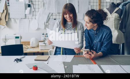 Attraktive junge Frauen in der Designbranche vergleichen Skizzen und sprechen darüber, während sie in einem modernen Schneidergeschäft arbeiten. Produktives Teamkonzept. Stockfoto