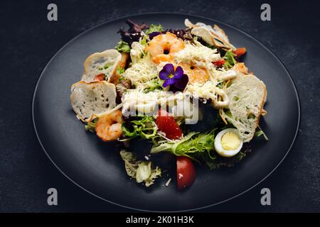 Caesar Salat mit Garnelen und Blumen, auf einem dunklen Teller, auf dunklem Beton Stockfoto