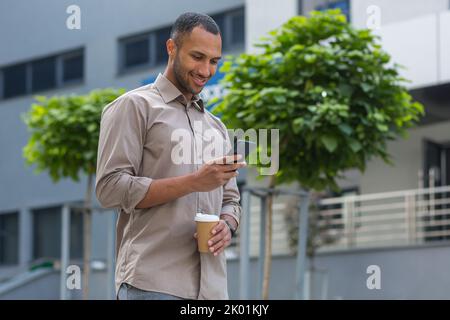 Ein afroamerikanischer Geschäftsmann, der lächelnd und glücklich vor das Bürogebäude geht, während einer Pause eine Tasse heißes Getränk in der Hand hält und das Telefon benutzt, Webseiten durchsucht und Nachrichten schreibt Stockfoto