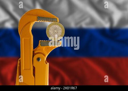 100 Kasachen-Tengenmünze, eingeklemmt in einen orangefarbenen Klempnerschlüssel auf dem Hintergrund der russischen Flagge. Vorderseite Stockfoto