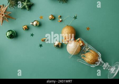 Weihnachten flach legen. Stilvolle weihnachts-Goldkugeln und Konfetti, die aus Champagner-Glas auf grünem Hintergrund gegossen werden. Grußkarte der kreativen Saison. Hap Stockfoto