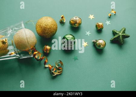 Frohes neues Jahr und frohe Weihnachten! Stilvolle weihnachts-Goldkugeln und Konfetti, die aus Champagner-Glas auf grünem Hintergrund gegossen werden. Kreative Saison gr Stockfoto