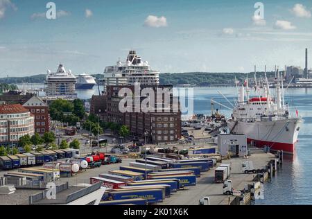 Kiel, Deutschland. 08. August 2022. Am Kreuzfahrtterminal Sartorikai des Kieler Hafens liegen zwei Kreuzschiffe hinter dem Gebäude der Unternehmen Sartori und Berger. Im Hintergrund kommt eine Fähre von Color Line Cruises an. Auf der rechten Seite befindet sich das Cap San Diego, das größte fahrtüchtige Museumsfrachtschiff der Welt. Davor stehen LKW-Anhänger auf einem Parkplatz. Quelle: Soeren Stache/dpa/Alamy Live News Stockfoto