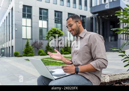 Junge afroamerikanische Studentin, die auf einer Bank außerhalb des Universitätscampus studiert, Mann, der einen Laptop für Videoanruf verwendet und sich mit dem Stift Notizen macht. Stockfoto