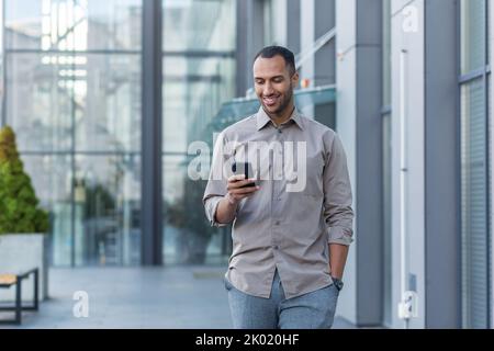 Erfolgreicher junger afroamerikanischer Mann, der vor dem Bürogebäude läuft, Ingenieur-Software-Entwickler-Programmierer lächelnd und glücklich mit Test-App am Telefon, glücklich zufrieden mit dem Ergebnis. Stockfoto