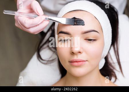 Kosmetologe, der schwarze Maske auf hübsches Frauengesicht mit schwarzen Handschuhen aufträgt, wunderschöne Frau im Spa, die Gesichtsbehandlungen hat Stockfoto