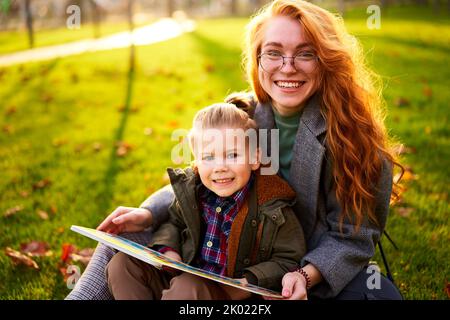 Rotschopf Frau liest Buch mit kleinen Jungen auf Gras im Stadtpark sitzen. Die junge Mutter unterrichtet ihren Sohn mit der ersten Klasse und macht Hausaufgaben am sonnigen Herbsttag Stockfoto