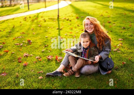 Rotschopf Frau liest Buch mit kleinen Jungen auf Gras im Stadtpark sitzen. Die junge Mutter unterrichtet ihren Sohn mit der ersten Klasse und macht Hausaufgaben am sonnigen Herbsttag Stockfoto