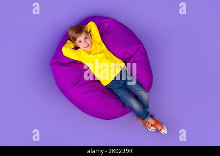 In voller Länge Foto oben Winkel Ansicht der hübschen Sohn tragen gelben Rollkragen sitzenden Sitzsack Arme hinter dem Kopf isoliert violetten Hintergrund Stockfoto