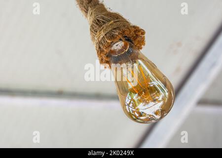 Eine alte Glühbirne hängt an einem Seil von der Decke. Eine wunderschöne dekorative Glühbirne wird aus nächster Nähe aufgenommen. Stockfoto