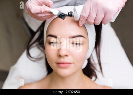 Kosmetologe, der schwarze Maske auf hübsches Frauengesicht mit schwarzen Handschuhen aufträgt, wunderschöne Frau im Spa, die Gesichtsbehandlungen hat Stockfoto