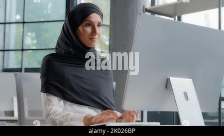 Junge arabische Geschäftsfrau in Hijab arbeitet am Computer lächelnd genießen Büroarbeit erfolgreiche Frau professionelle Manager Firma Mitarbeiter entwickelt Stockfoto