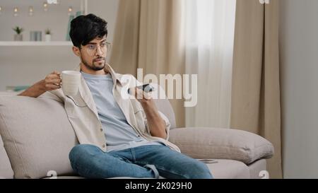 Arabischer Mann bärtiger Kerl mit Brille, die zu Hause auf der Couch sitzt, Tee trinkt, Kaffee schaut lustige Film-Meisterschaft Sport-Spiel-TV jubelt Stockfoto